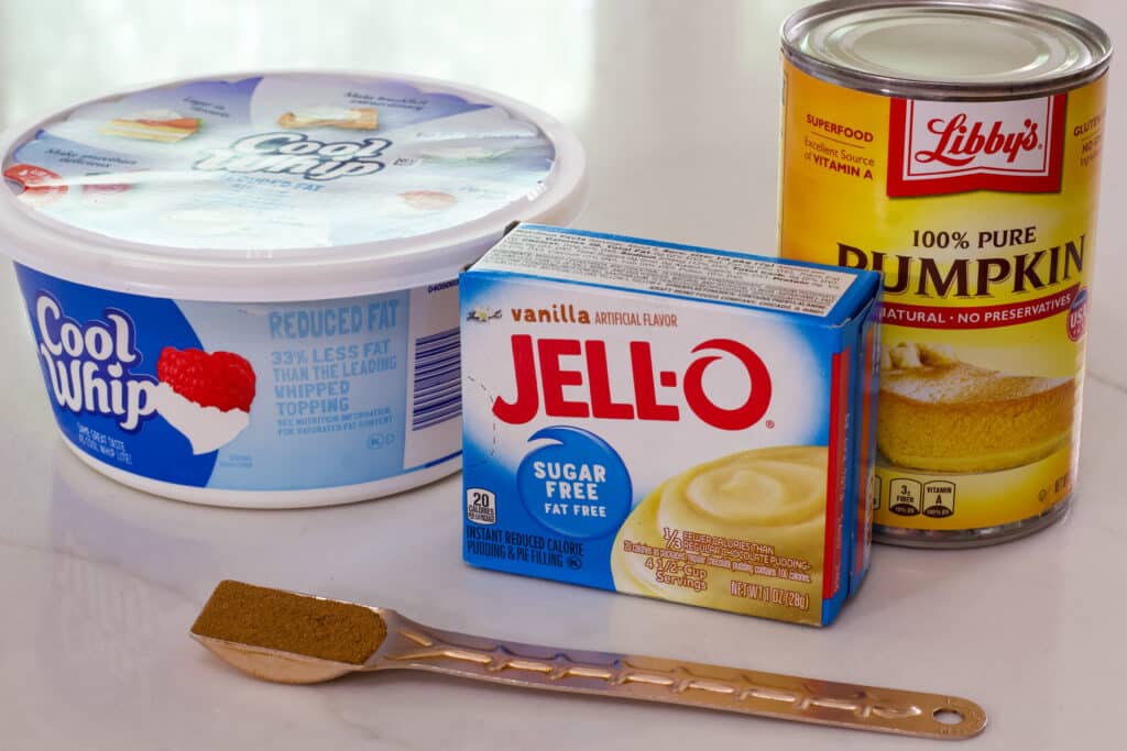 The container of cool whip. box of pudding, can of pumpkin and teaspoon of pumpkin pie spice on the counter ready to be used to make the recipe.