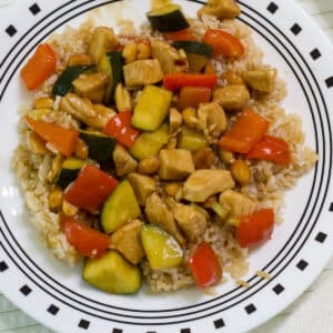 A white plate with a serving of kung pao chicken on a bed of brown rice.