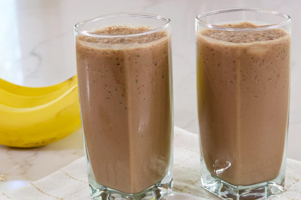 Two glasses full of Easy & Healthy Chocolate Coffee Smoothie with a bunch of bananas sitting next to them.