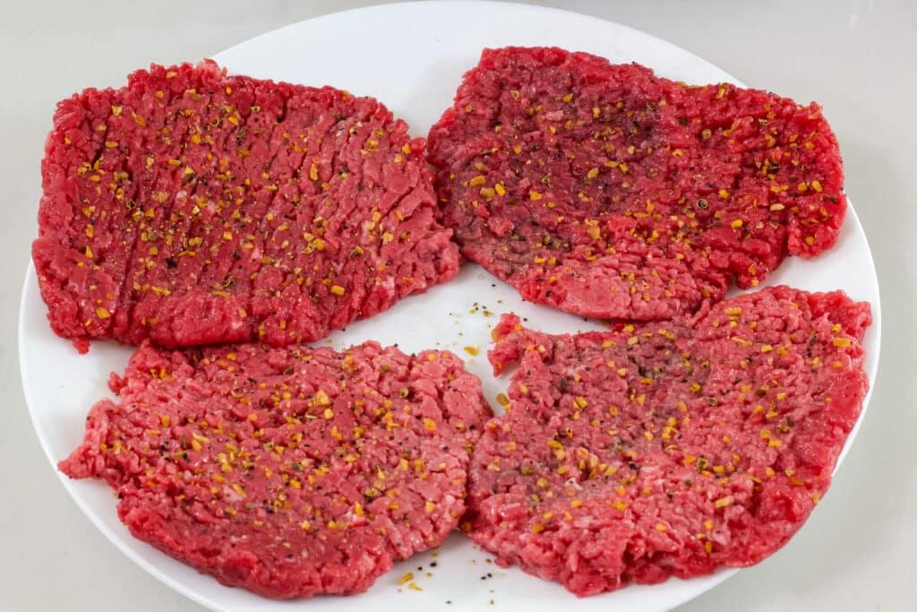 Four uncooked cube steaks on a white plate, they are seasoned with Montreal steak seasoning.