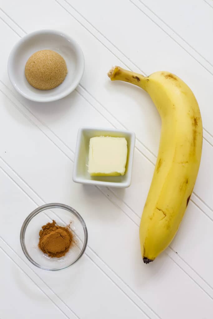 One banana in the peel and the butter, brown sugar, and cinnamon measured out in small bowls on a white background.