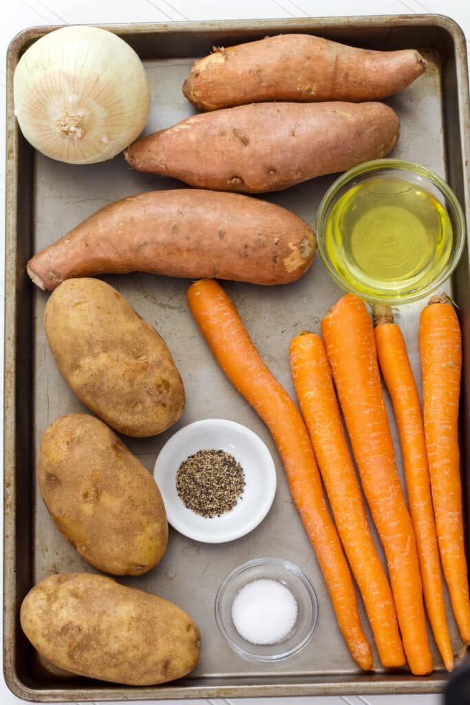 A rimmed baking sheet with 3 sweet potatoes, 3 russet potatoes, 5 carrots, one onion, a small bowl of olive oil, a small bowl of pepper and a small bowl of salt.