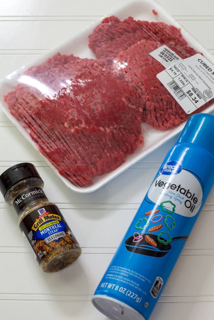 A package of cube steaks, bottle of montreal steak seasoning and can of nonstick cooking spray on a white table.