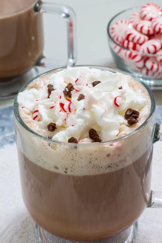 one mug of peppermint mocha in the foreground and another mug of coffee and a small bowl of peppermint candies in the background.