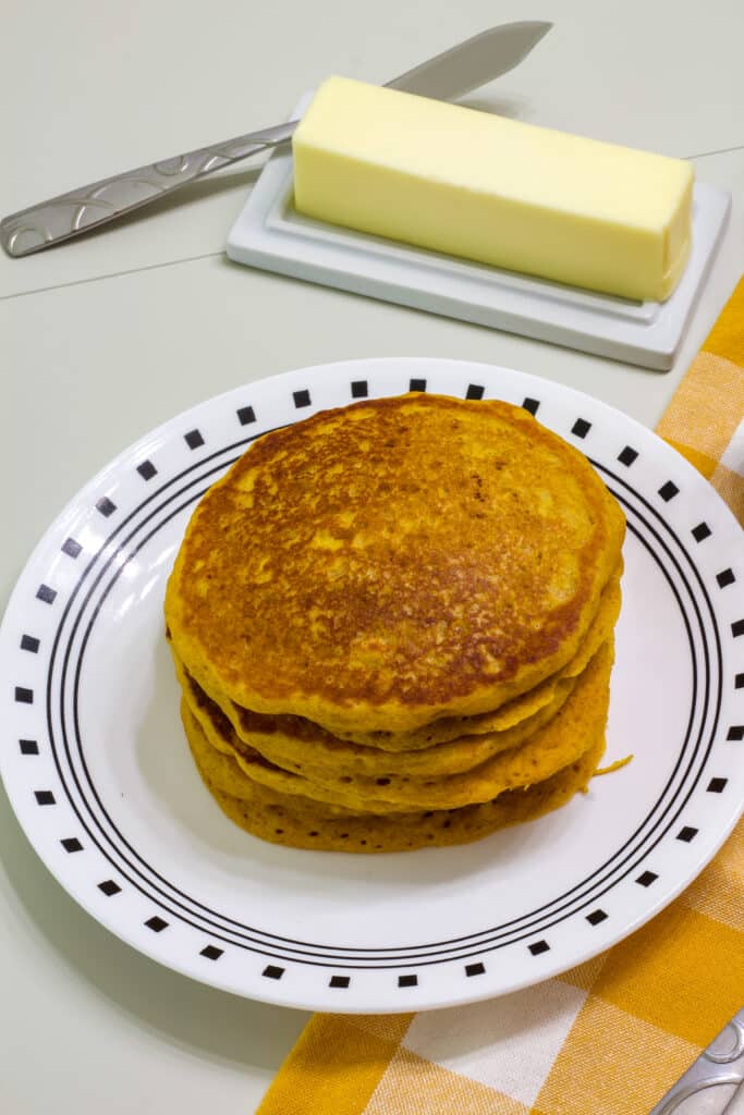 Four pumpkin pancakes on a plate and a stick of butter on a dish.