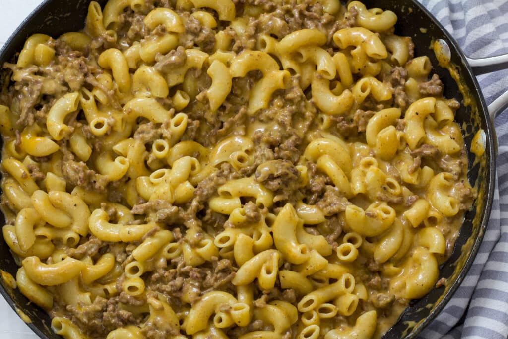 A skillet full of homemade hamburger helper cheeseburger macaroni and a grey and white striped tea towel next to it.