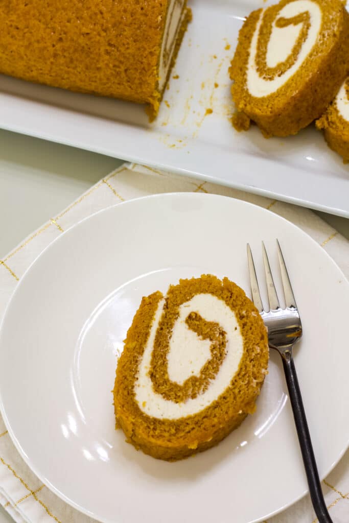 One slice of pumpkin roll on a plate in the foreground and the remaining pumpkin roll in the background.