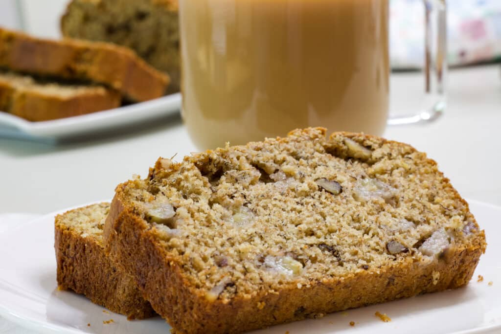 A couple of slices of banana bread in the foreground and a cup of coffee and the remaining loaf in the background.