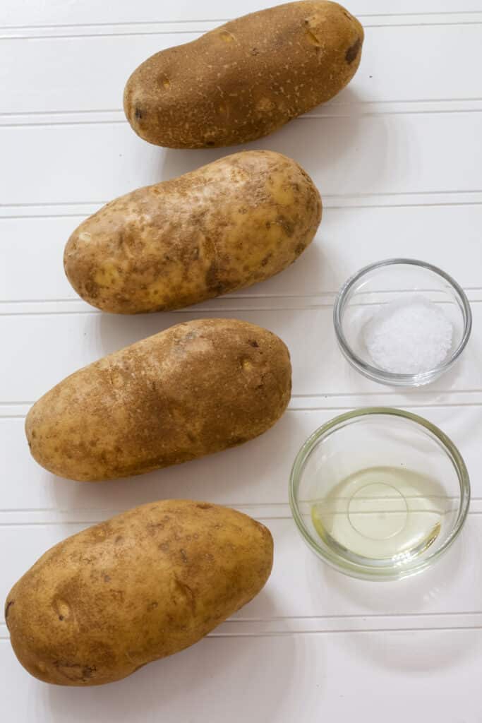 Four potatoes, a small bowl of canola oil and a small bowl of kosher salt on a white background.