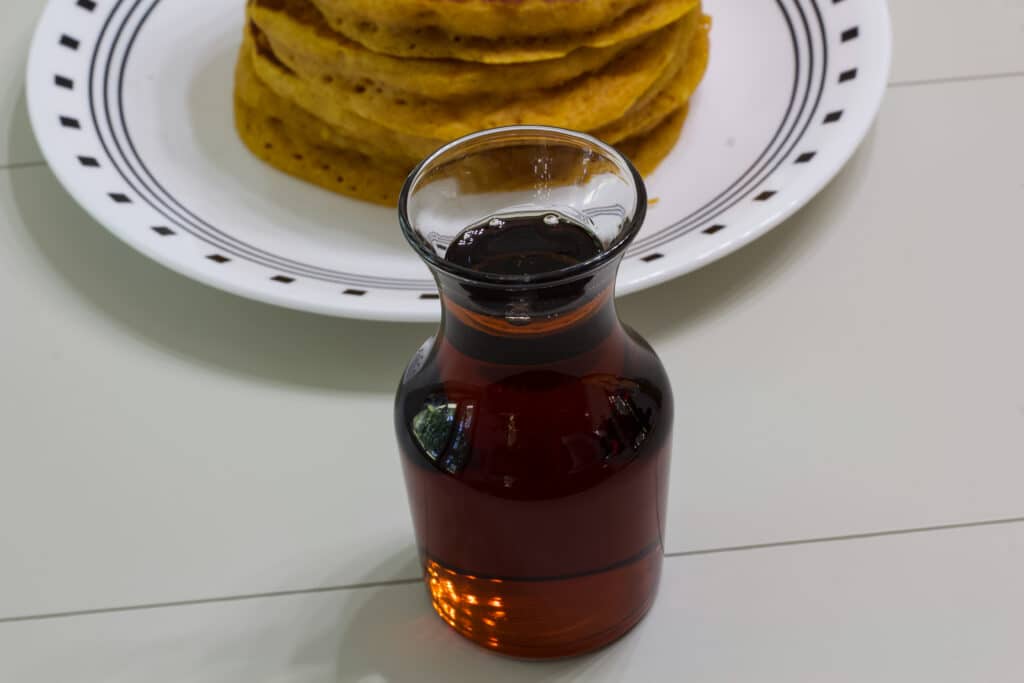 A small bottle of 2 Ingredient Copycat IHOP Butter Pecan Syrup in front of a plate of pumpkin pancakes.