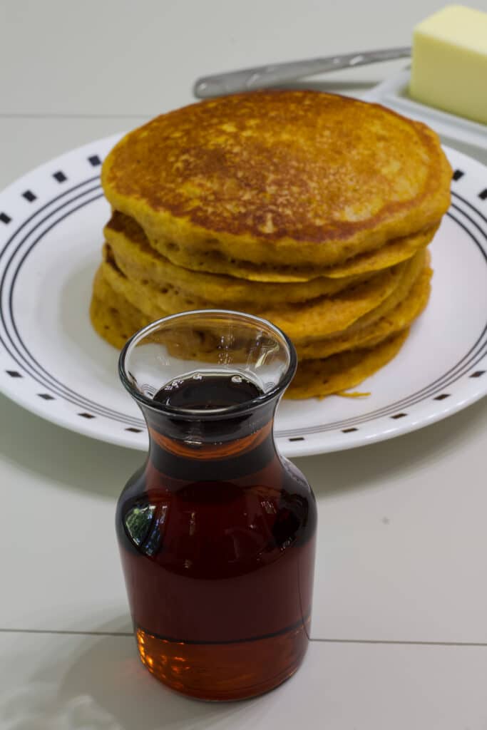 A small bottle of 2 Ingredient Copycat IHOP Butter Pecan Syrup in the foreground and a plate of 4 pumpkin pancakes in the background.