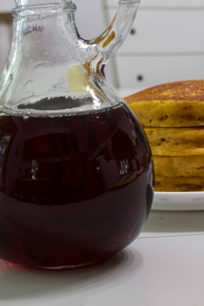 Close up of a small bottle of 2 Ingredient Copycat IHOP Butter Pecan Syrup and a portion of a stack of pumpkin pancakes is visible.