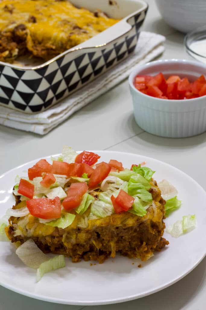 A serving of taco salad casserole on a plate topped with lettuce and tomatoes in the foreground and the rest of the casserole in the background.