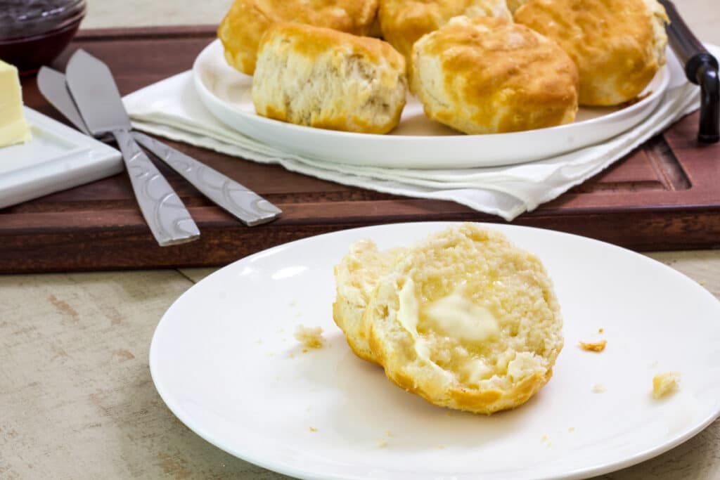 A sliced biscuit with butter on it in the foreground and a plate with many biscuits in it in the background.