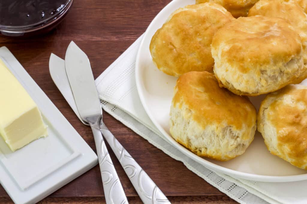 A stick of butter, bowl of jelly, two butter knives and a plate of biscuits on a wooden serving tray.