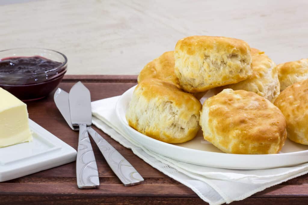 Side view of a plate ob biscuits, small bowl of jelly, 2 butter knives and a stick of butter on a serving tray.