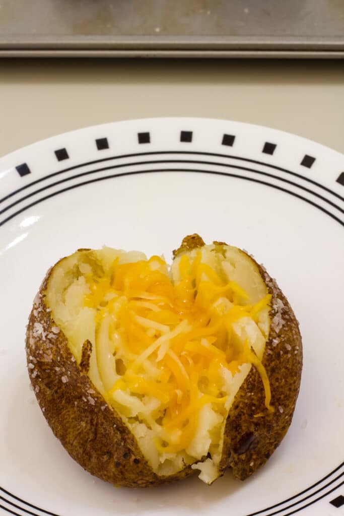 One oven baked potato that is topped with shredded cheese that has melted a little bit.