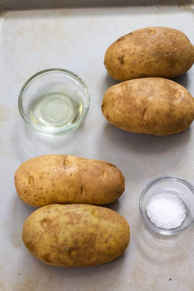 Four raw potatoes, a small bowl of canola oil and a small bowl of salt on a rimmed baking sheet.