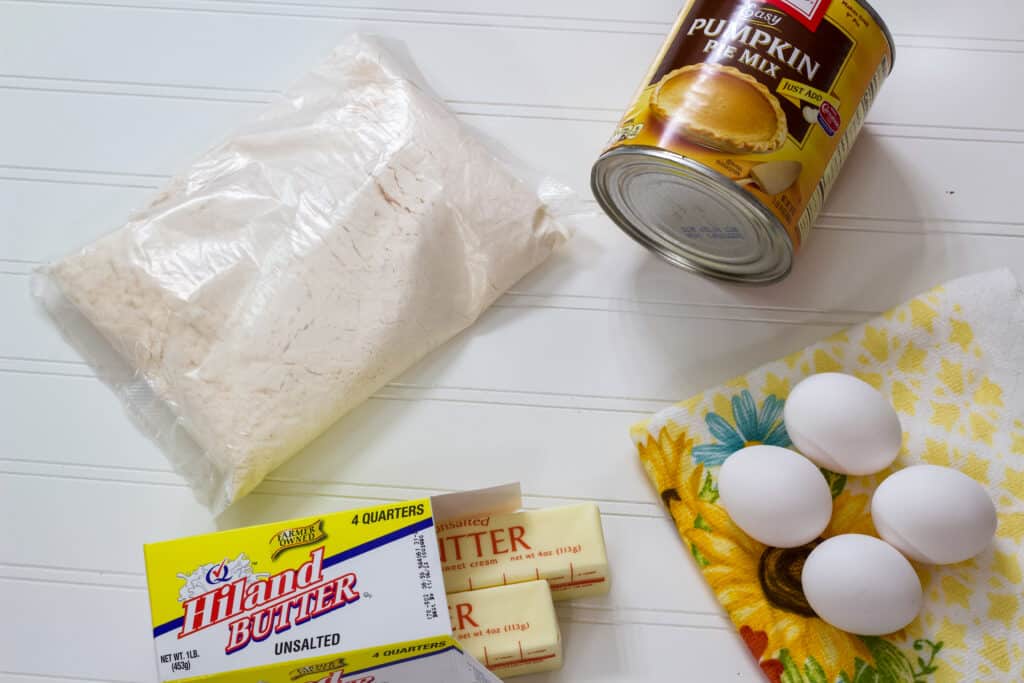 The dry cake mix, can of pumpkin pie mix, 4 eggs and 2 sticks of butter laying on a white table.