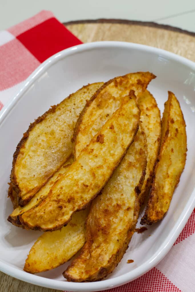 Several cooked potato wedges on a white plate this is sitting on a white and red checkered napkin.