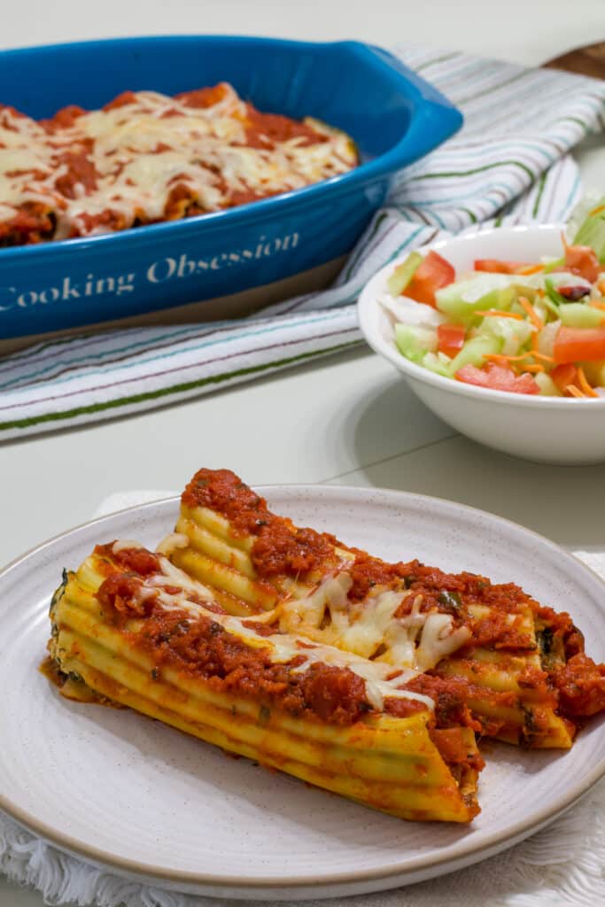 Two manicotti on a plate in the foreground and the casserole dish full of manicotti and a small salad in the background.