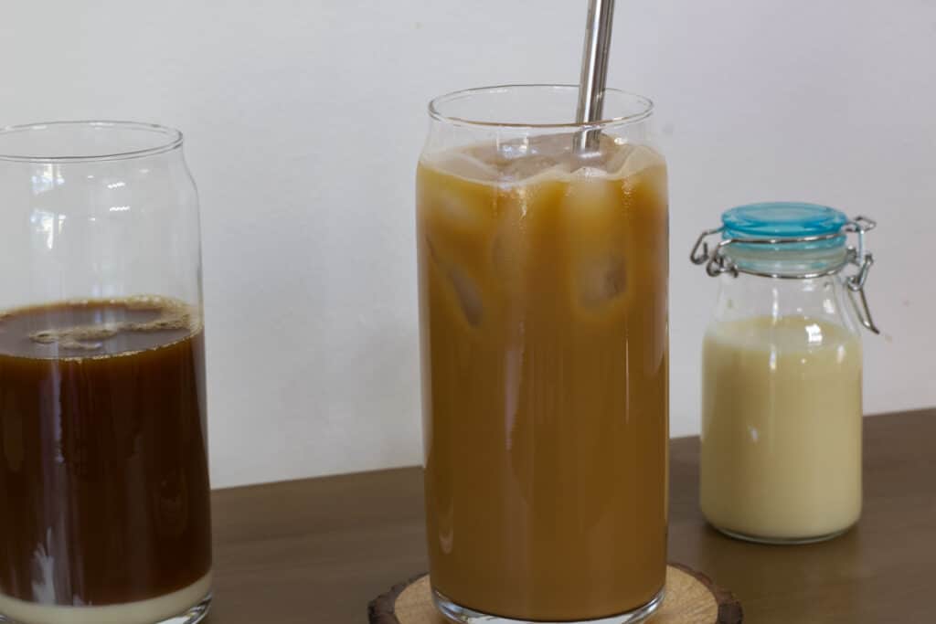 One mixed glass of ice coffee, one with just the condensed milk and black coffee in it unmixed and a small bottle of sweetened condensed milk.