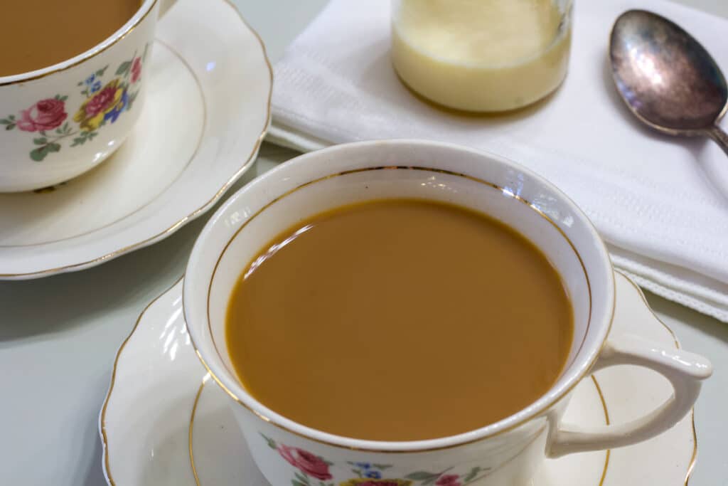 One hot cup of coffee with Sweetened Condensed Milk, there is a silver spoon and a small bottle of milk next to it.