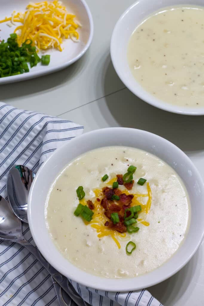 A bowl of soup topped with cheese, scallions and bacon and one bowl without toppings, there are toppings on a plate too.