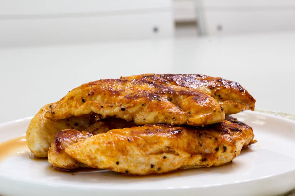 Five cooked chicken tenders, two on top of three, on a white plate.