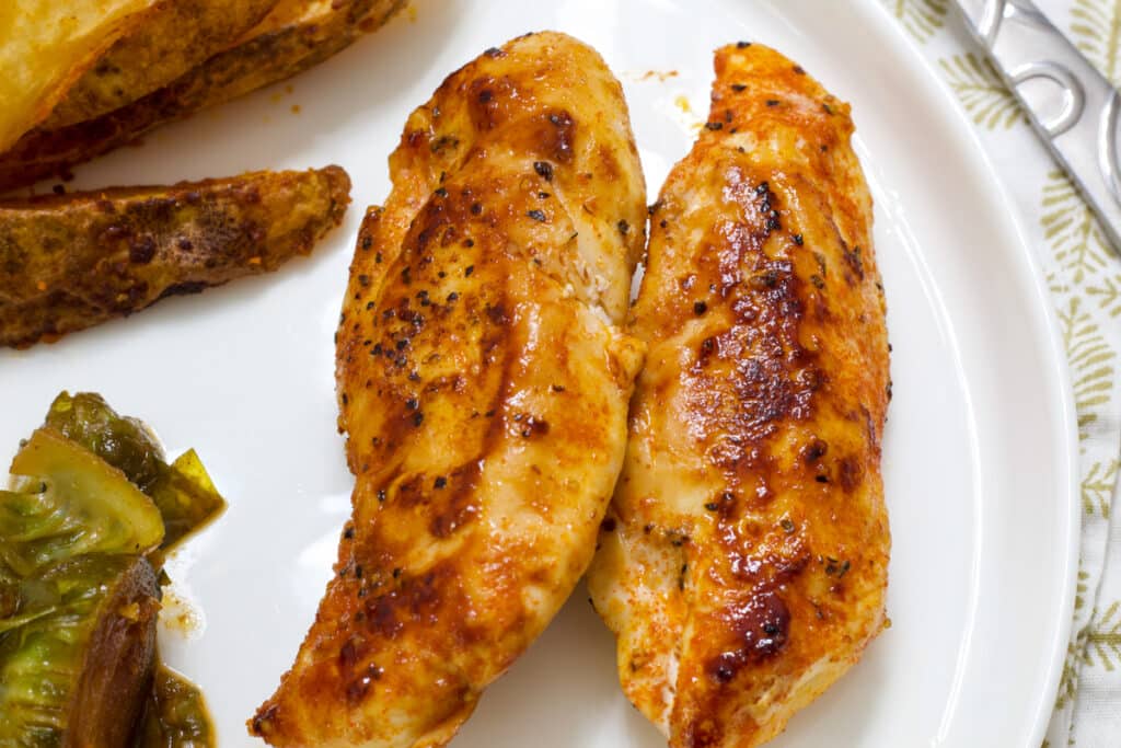 Two cooked chicken tenders on a white plate, you can barely see the potato wedges and brussels sprouts on the plate next to them.