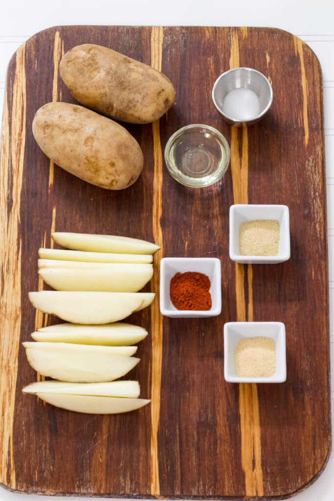 The potatoes, 2 uncut and 1 cut, oil, and seasonings needed to make Easy 20 Minute Air Fryer Potato Wedges Recipe on a wooded cutting board.