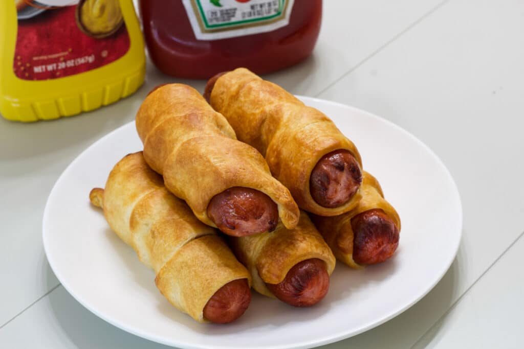 Five pigs in a blanket on a white plate with a bottle of ketchup and mustard in the background.