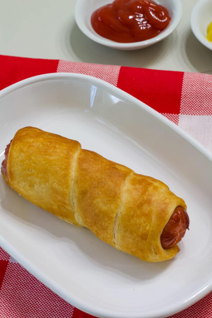 One pig in a blanket on a white plate with small bowls of ketchup and mustard in the background.