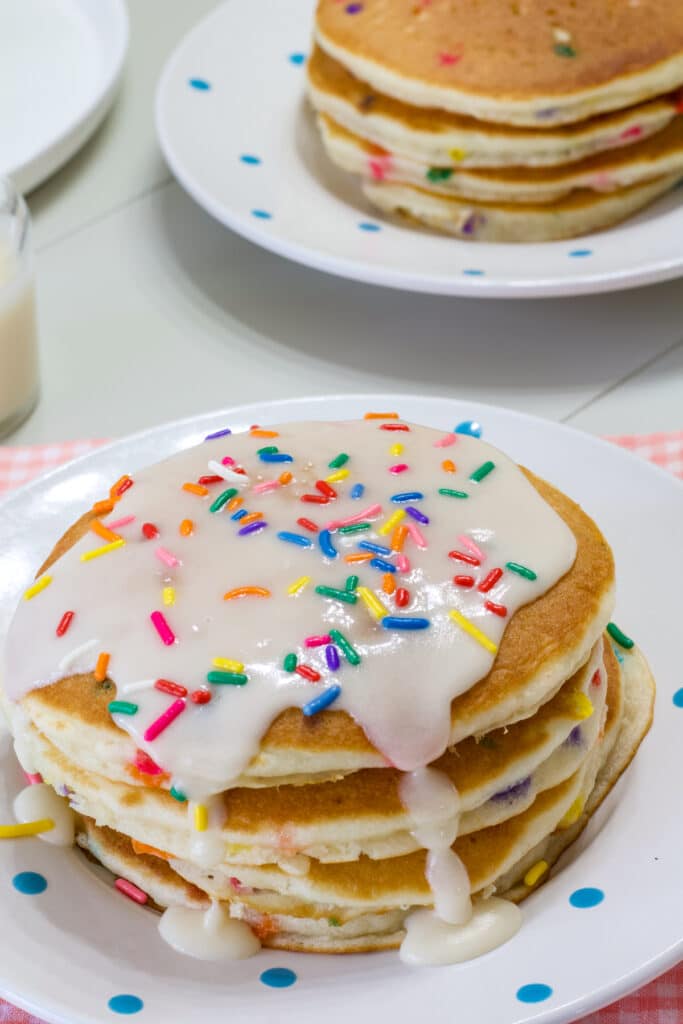 A stack of four funfetti cake mix pancakes with vanilla icing on them on a white plate with blue dots.