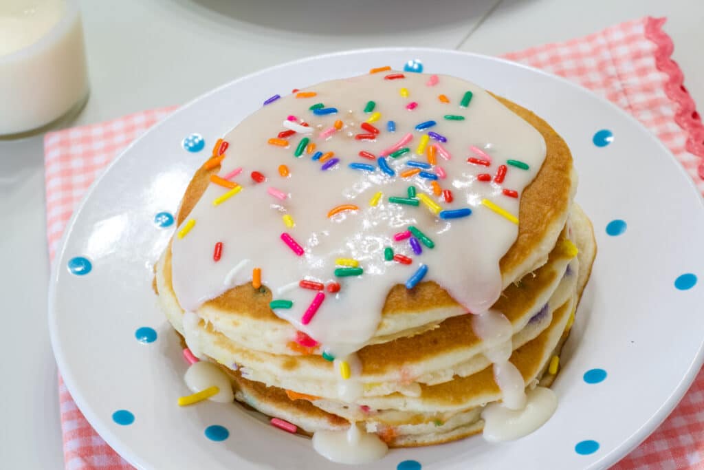 A stack of frosted pancakes on a plate that is sitting on a pink and white checkered napkin.
