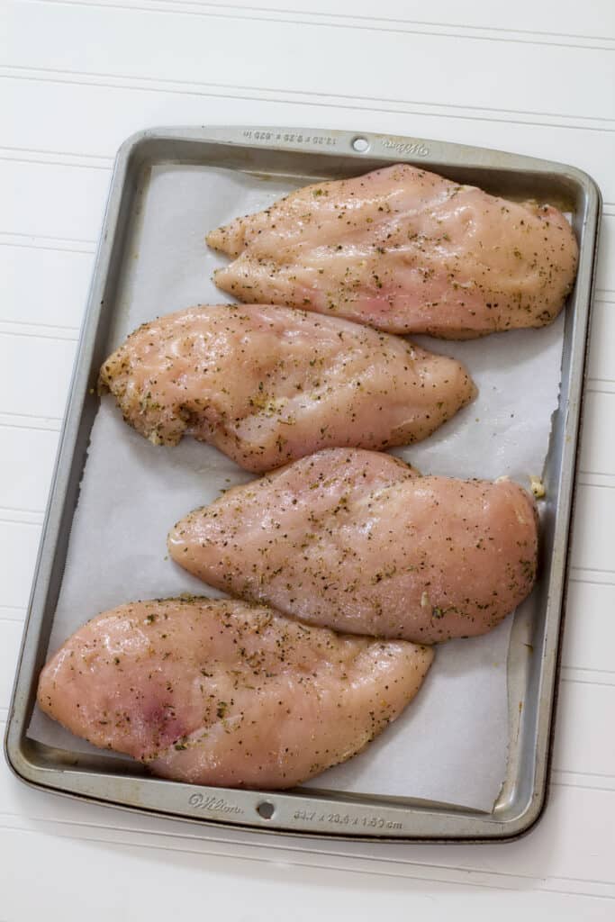 Four raw chicken breasts that have been seasoned on a sheet pan ready to go into the oven.