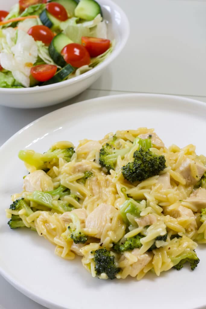 A serving of Cheesy Chicken & Broccoli Orzo in the foreground and a bowl of salad in the background.