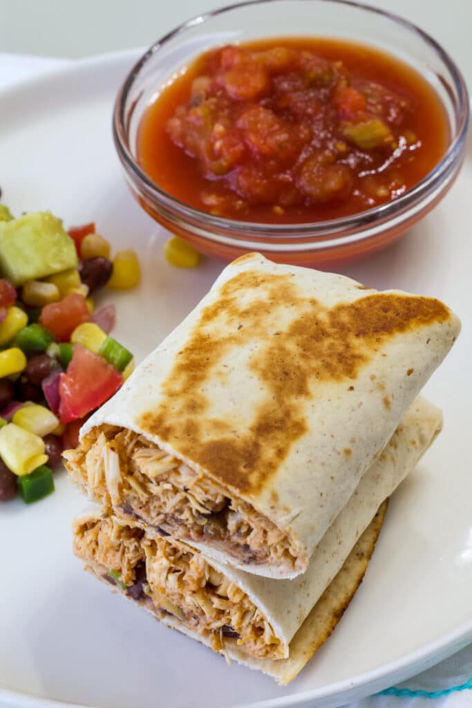 One burrito cut in half on a white plate with a small bowl of salsa and serving of corn salad.