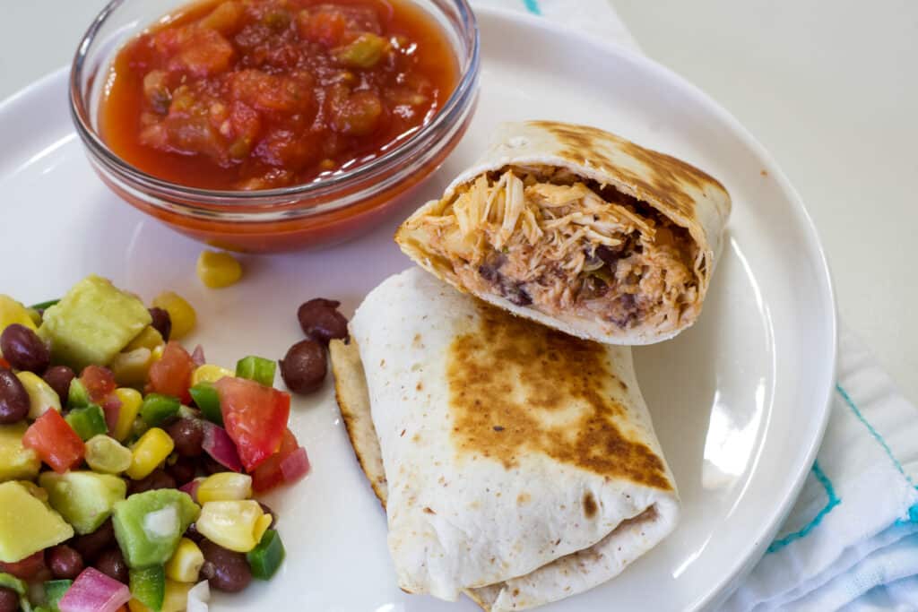 A burrito on a plate so the inside is visible, there is a small bowl of salsa and some corn salad on the plate too.