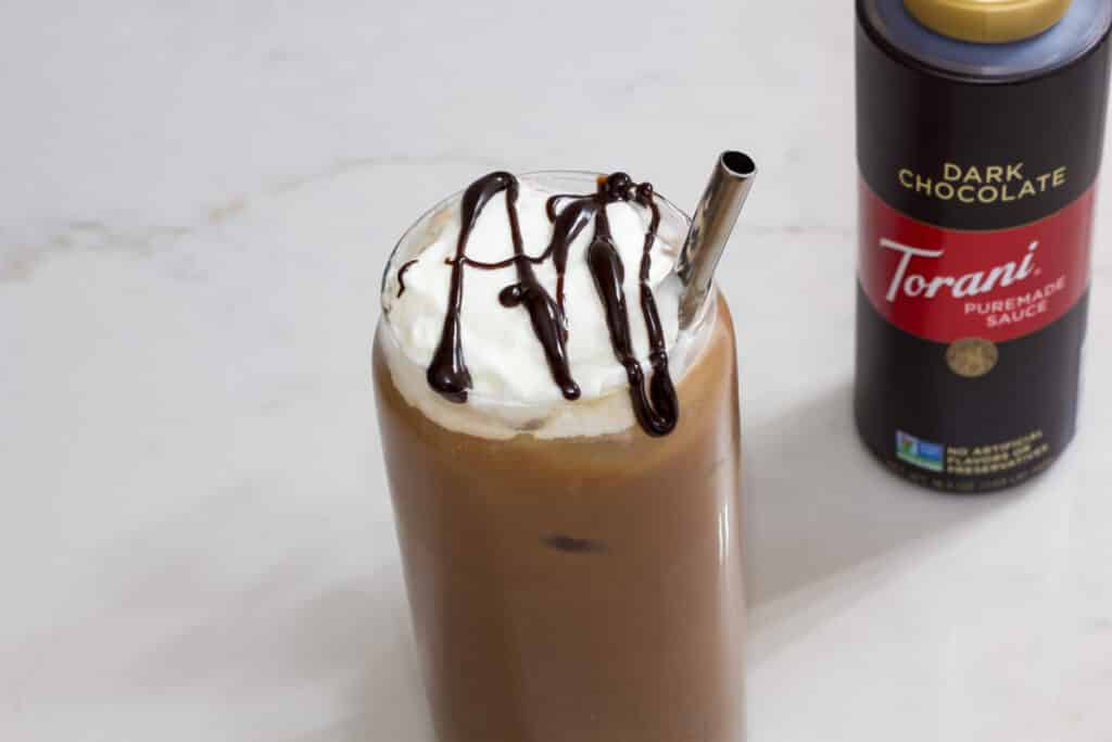 The top of the glass of iced mocha coffee and the bottle of Torani dark chocolate sauce sitting on the counter.