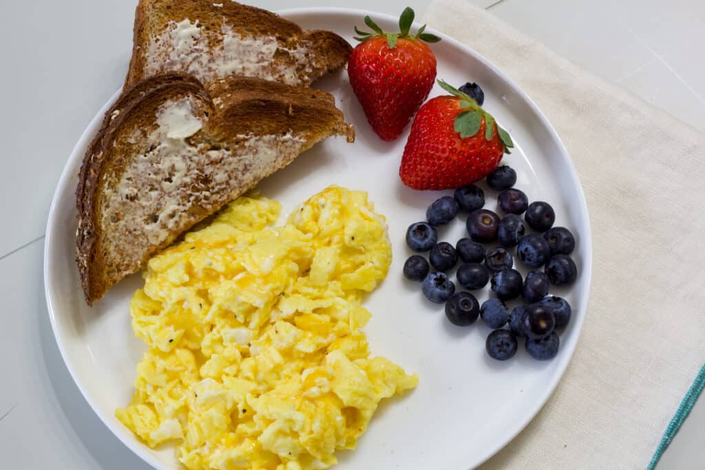 A white plate with scrambled eggs with cheese, two pieces of wheat toast, two strawberries and some blueberries.