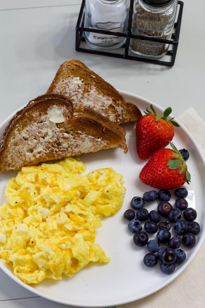 Scrambled eggs with cheese on a white plate with toast and fresh strawberries and blueberries.