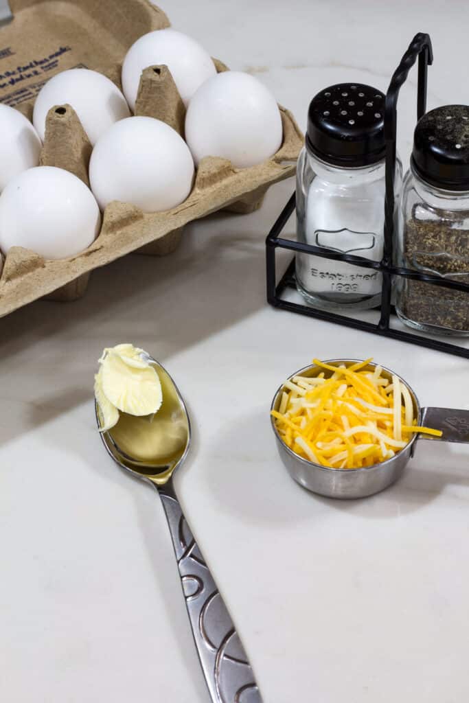 A carton of a dozen eggs, salt and pepper shakers, a spoon half full of butter and ¼ cup of shredded cheese.