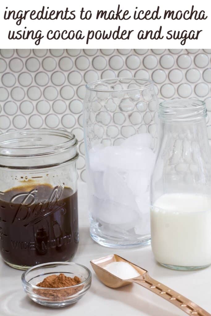 A mason jar of coffee, bottle of milk, glass of ice, small bowl of cocoa powder and a tablespoon with some sugar in it.