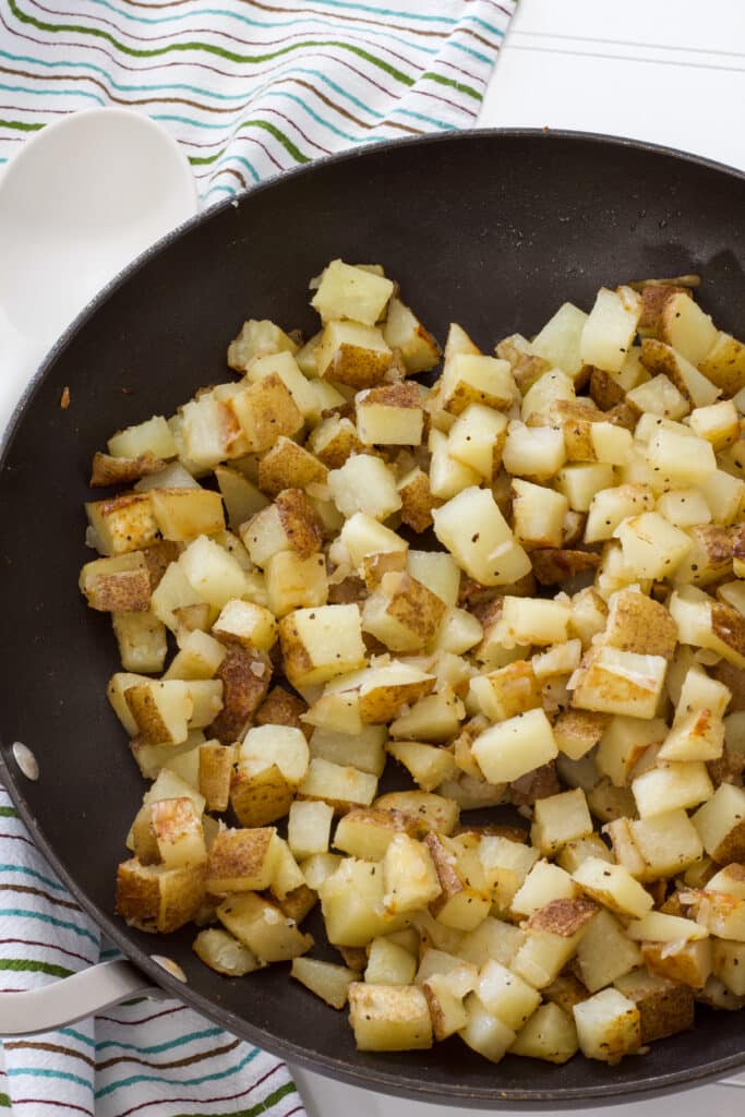 A skillet full of cooked Homemade Pan-Fried Potatoes and a turquoise and brown striped tea towel next to it.