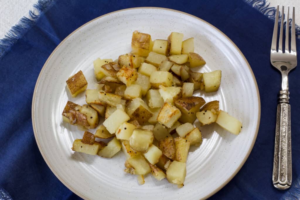 A serving of fried potatoes on a plate sitting on a blue napkin with a fork sitting next to the plate.