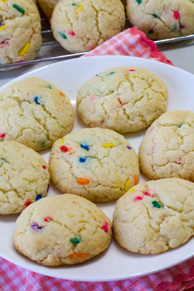 Several baked cookies on a white plate with cookies on a cooling rack in the background.