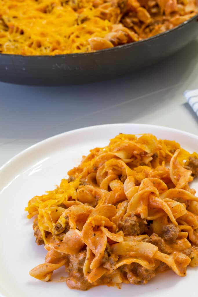 One serving of hamburger casserole on a plate in the foreground and the skillet in the background.