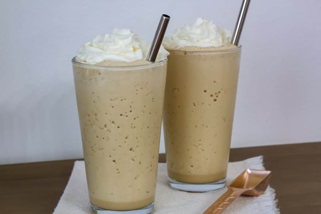 Two glasses of vanilla frappuccino sitting on a napkin with a copper tablespoon sitting next to them.