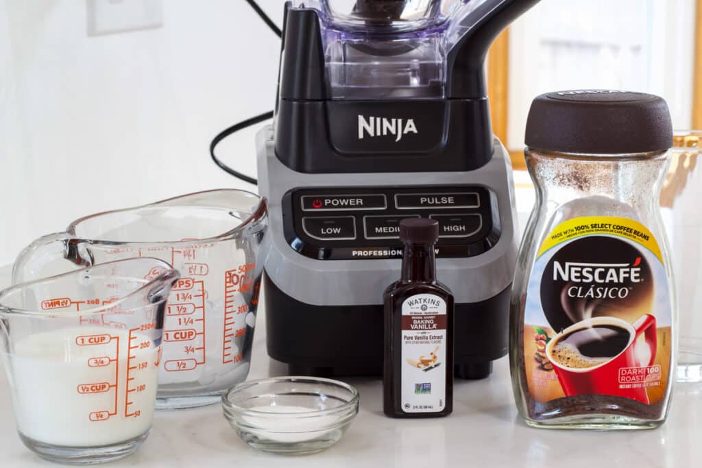 The instant coffee, milk, ice, sugar and vanilla needed to make a vanilla frappuccino sitting on the counter in front of a ninja blender.